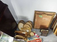 Lot of various vintage and antique picture frames stacked on carpeted floor, some gold-toned with ornate designs, some oval and some rectangular, arranged against a white wall.