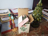 Green ceramic Christmas tree, box of vintage Christmas light bulbs, and two vintage candle lights adjacent on floor with stacks of papers in background