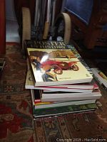 Stack of art books on patterned carpet with large handles of a chair in background. Shows the overall lot and book arrangement.