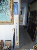 Tall vintage Da-Lite slide and movie projection screen box beside a black umbrella with wooden handle with framed art in background.