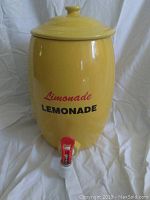Front view of the yellow ceramic lemonade dispenser jug with lid and visible red and white spigot and lettering on the front.