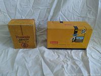 Two vintage Kodak Brownie camera boxes placed on a white cloth background, showing the front and side of the boxes.