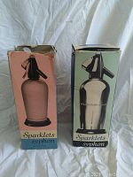 Two Sparklets syphon seltzer bottles in their original boxes, one pink-label box and one green-label box, displayed side by side.