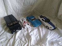 Four diecast model cars arranged side by side on white cloth including black off-road vehicle, white futuristic car, blue classic car, and white sports car with blue stripes.