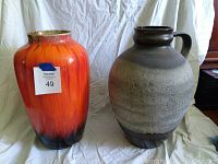 Two large pottery vases side by side on a white cloth background. One with a bright orange-red glaze, the other with grey-black handle and textured glaze.