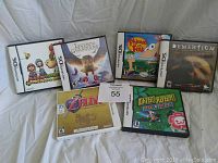 Six Nintendo DS games in their original cases arranged on a white cloth background. Visible titles include Zelda, Mario, Legend of the Guardians, Phineas and Ferb, Dementium, and Chibi-Robo.