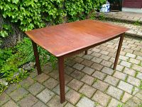 Side angle view of the teak table showing the rectangular top and one leaf installed, standing on a stone patio with green foliage background.