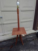 Full view of the mid century modern teak floor table lamp showing the full height, central column, square tabletop, tripod base, and bulb socket with bulb installed.