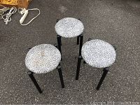 Three grey black metal stools with patterned round seats, one with brown stain on seat