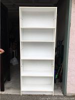 Front view of the tall white shelving unit showing four shelves and adjustable holes on the side panels.