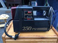 Black Superex air tire inflator with 250 PSI capacity, placed on a wooden surface with attached power cord and air hose.