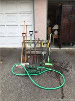 Photo showing full assortment of garden tools including shovels, garden pick, rakes, hand tools, brushes, and hoses arranged in front of garage door.