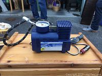 Side view of blue Pit Stop 12V air tire inflator compressor with gauge and attached hose lying on wooden surface.