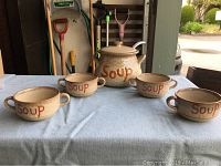 Five-piece soup set on a table including one large soup bowl with lid and ladle and four smaller soup bowls with handles, all beige ceramic with 'Soup' printed in orange.
