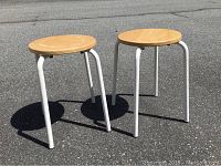 Photo showing two small identical stools with round wooden seats and white metal legs, placed outside on asphalt.
