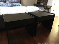 Photo of two black rectangular bedside tables side by side on wooden floor, showing overall condition and size relative to bed.