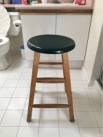Front view of a wooden stool with a round green seat and natural wood legs with three horizontal supports.