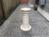 Full view of the cream coloured glazed ceramic plant stand standing upright outside on a concrete surface next to a house wall and a fence.