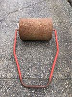 Metal lawn roller showing rusty roller and red metal handle lying on concrete ground from angled front view.