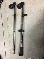 Close-up photo showing two stainless steel Scotty downrigger booms side by side on a concrete floor, detailing the pulleys, clamps, and ends of each boom