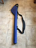 Full length view of blue fishing rod protection case lying flat on tile floor with shoulder strap extended.
