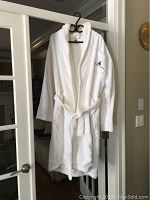 Front view of white terry cloth bathrobe hanging on a door, showing shawl collar, waist belt, and sailboat embroidered patch pocket on the chest.