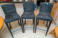 Three black fabric bar stools with wooden legs arranged side by side on a tiled floor.