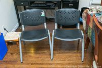 Front view of two black plastic chairs with gray metal legs, showing seat and perforated backrest detail.