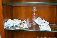 Glass shelf inside wood cabinet with candles, glass candle stick holders, pug figurines, serving knife, cloth napkins, and a glass decanter bottle with text