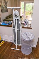 Folded ironing board standing against a counter, laundry hamper collapsed and an iron placed on the counter