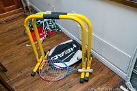 Yellow total body strengthener exercise equipment with tennis racket and HEAD racket carry bag on wooden floor near wall vent.