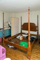 Full view of the wooden four-poster bed frame with headboard, footboard, and four tall posts in a room with wood flooring and surrounding furniture.