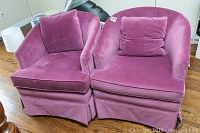Front view of two purple cloth upholstered chairs with loose back cushions and skirted bottoms.