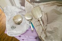 Three sterling silver vanity items displayed on tissue paper: a round jewelry box with feet, a hand mirror, and a hairbrush, all engraved and monogrammed.