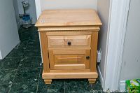 Front view of light wood end table with drawer and door, showing black knobs and carved decorative trim.