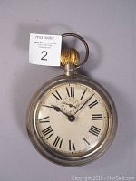 Front view of the oversized pocket watch style alarm clock showing white metal case, Roman numeral dial, black hands, and subdial for alarm at center bottom.
