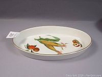 Side angled view of large oval ceramic baking pan showing colorful corn and fruit motifs with gold rim on white ceramic.