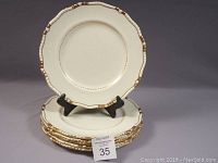 Six cream-colored dinner plates with gold encrusted scalloped edges stacked with one displayed upright on a stand