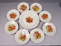Eight dessert plates and one large serving tray arranged with visible fruit and nut designs and gold rims