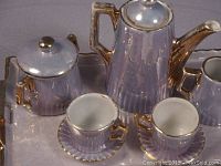 Close-up image of antique Victorian doll tea set pieces arranged on tray highlighting teapot, sugar bowl, cream pitcher, cups and saucers with visible gold accents and purple finish