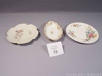 Three ceramic plates: Limoges Elite Works small oval platter with embossed floral details, Royal Doulton Arcadia floral round plate, and ruffled rim plate with floral pattern and small chip on rim.