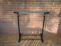 Assembled garment rack with black metal base and vertical poles, chrome adjustable hanging bar, standing indoors against brick wall.