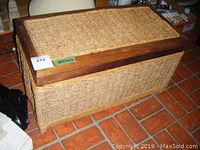 Photo showing woven rectangular chest with hinged lid, wooden frame on top, and woven sides standing on tiled floor.