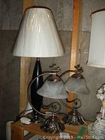Three table lamps on a wooden shelf against wall; two smaller twin-shade lamps with ornate metal bases and curved arms, one tall black lamp with off-white shade.