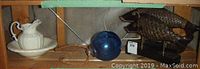 Photo showing small white ceramic wash basin with water jug on a wooden shelf next to blue glass ball and decorative fish.