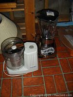 Proctor-Silex food processor and Oster blender together on a tiled floor near a wooden chair, showing overall appliances.