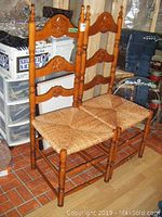 Pair of wooden ladder back rush seat chairs with carved sunburst designs on the back rails. Chairs show minor scuffs. Positioned on reddish tile floor with miscellaneous items in background.