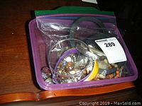 Photo of various costume jewelry pieces inside a clear plastic bag with a purple container underneath, showing bracelets, earrings, and necklaces mixed together.