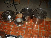 Photo showing large canning pots, small copper pots and frying pan with lids arranged on tiled floor.