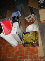 Photo of assorted kitchen items on floor with boxes, plastic containers, red vinyl table cloth, small tins, and plates visible.